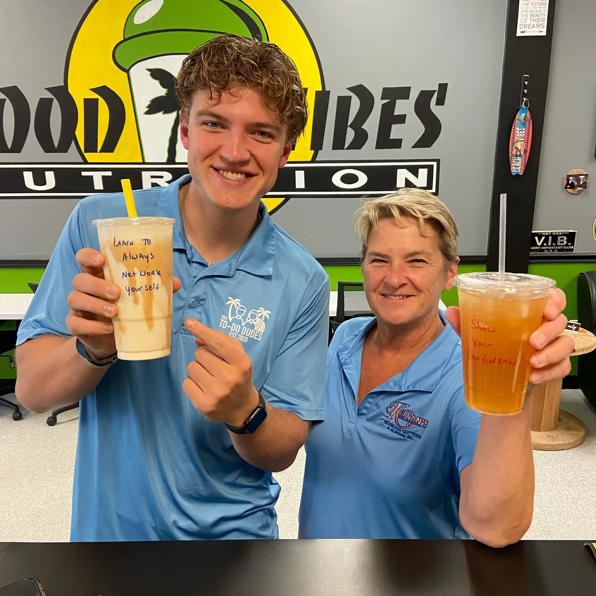 Two customers hold up their drinks in front of the Good Vibes wall.