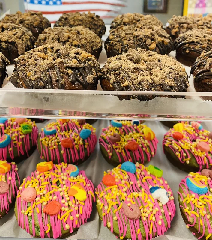 A display case of protein donuts: chocolate with crushed cookies on top, and bright ones with pink icing and cereal pieces.