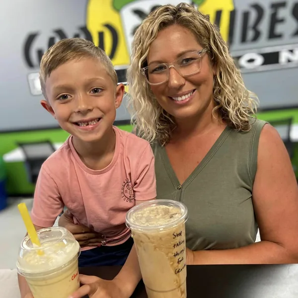 A smiling woman and a young boy at a table, each holding a cold drink, in front of the colorful mural.