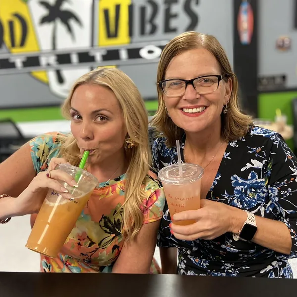 Two smiling women in floral dresses sit side by side with iced drinks in front of the Good Vibes Nutrition wall.