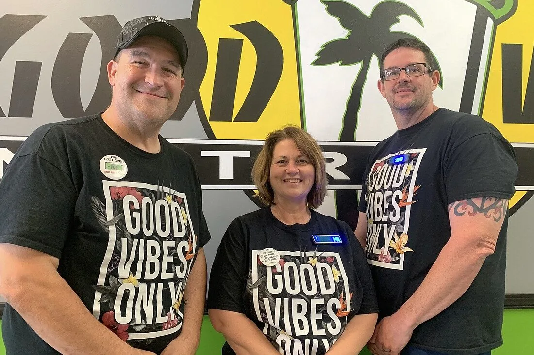 Three people in black 'Good Vibes Only' shirts in front of the club's mural.