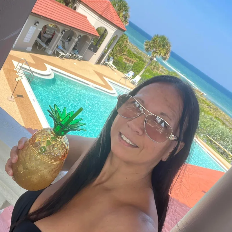 A customer in sunglasses holds a gold pineapple cup by a pool with the ocean behind her.